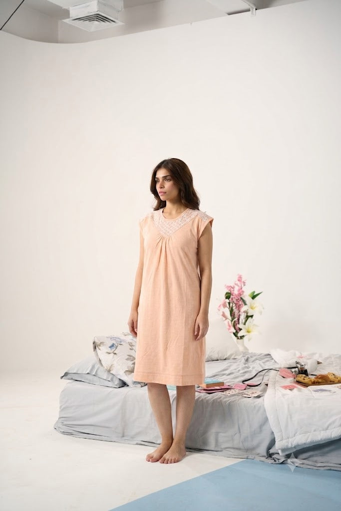 A woman wearing a light peach-colored sleeveless nightgown stands barefoot in a minimalist white studio setting with a bed and flowers in the background.